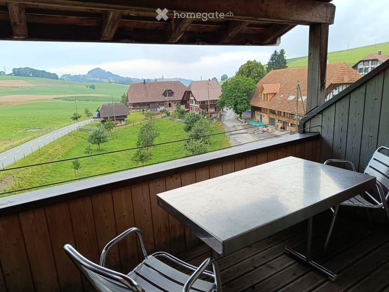 Balcony with wooden railing, metal table, and chairs, overlooking a scenic countryside with houses and green fields.