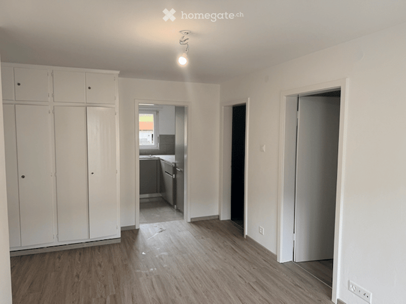 Empty room with wooden flooring, white walls, built-in wardrobes, and a view into an adjacent kitchen area.