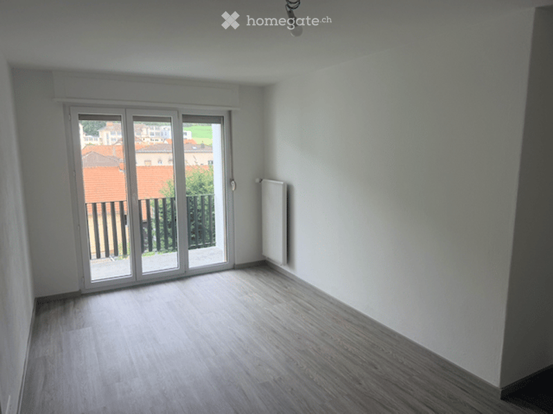 Empty room with wooden flooring, white walls, large sliding glass door leading to a balcony, radiator on the wall, and a view of the outdoor area.