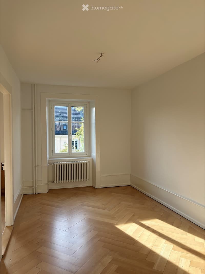 Empty room with light wooden herringbone flooring, white walls, and a large window with white frame showing a view of neighboring buildings. A white radiator is placed below the window.