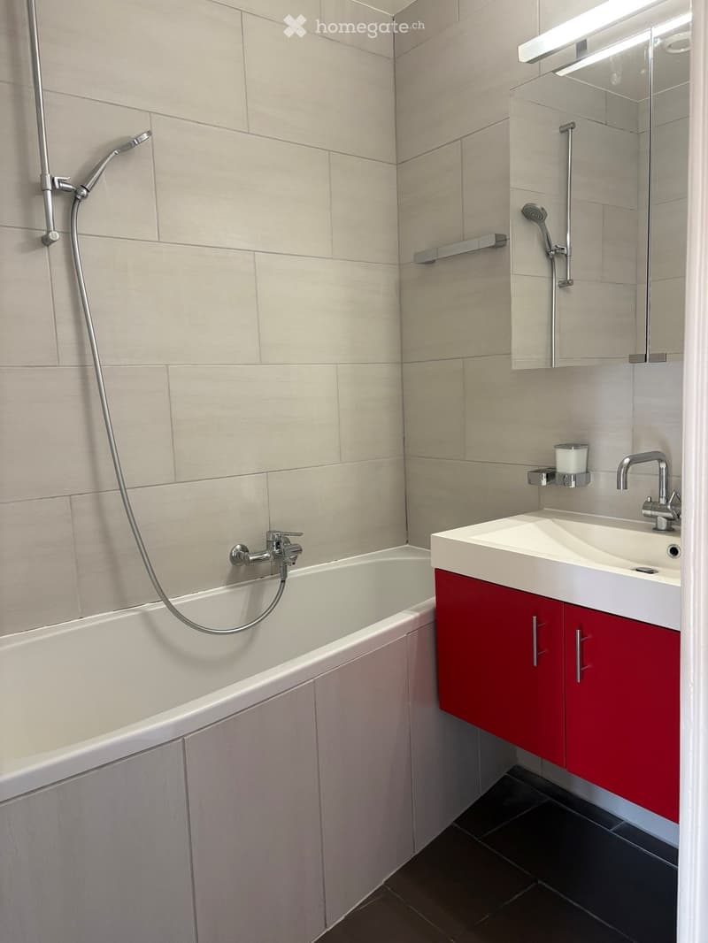 Modern bathroom with a white bathtub, red cabinet, white sink, mirror, and showerhead.