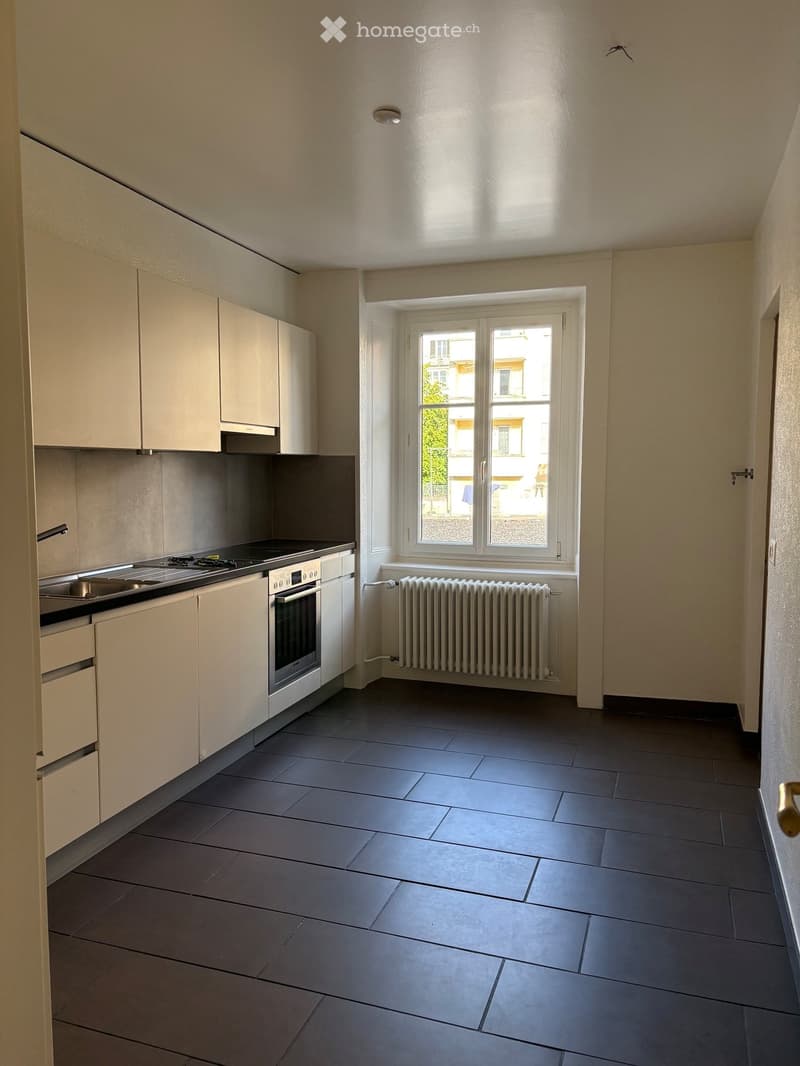 Modern kitchen with light-colored cabinets, a stove, oven, sink, and radiator under a large window.