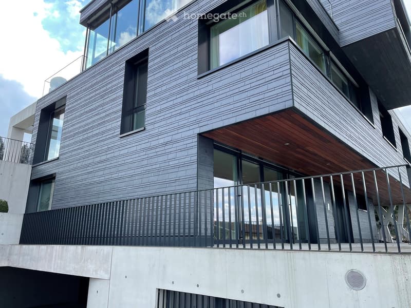 Modern multi-story building with grey brick facade, large windows, and a wooden balcony with metal railing.