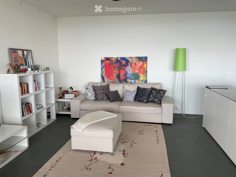Modern living room with a light-colored sectional sofa, ottoman, bookshelf, small table, floor lamp, and artwork on the wall.
