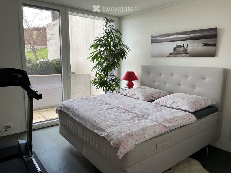 Modern bedroom with a large double bed, floral bedding, a red table lamp, a picture frame on the wall, and an exercise bike near the window.