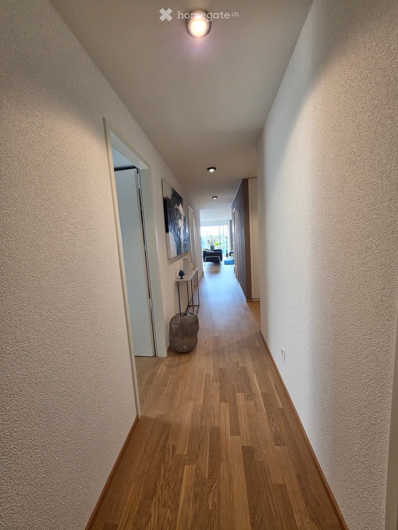 A modern hallway with wooden flooring, white walls, and recessed lighting. Features a small table with a decorative item and a woven basket on the floor.