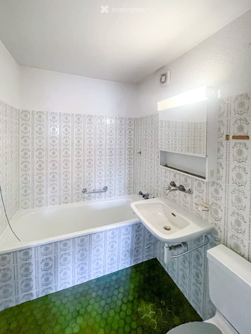 Bathroom with a bathtub, sink, toilet, and mirror. The walls are tiled with a decorative pattern, and the floor has a green artificial grass covering.