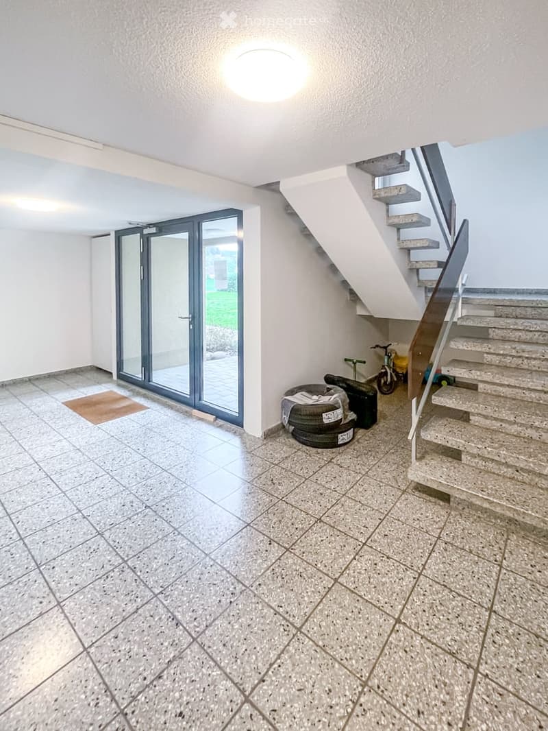 A modern, clean hallway with a staircase leading upwards, tiled flooring, and a large glass door opening to an outdoor area. The space includes a small rug near the door and some stored items, including tires and a bicycle, against the wall.