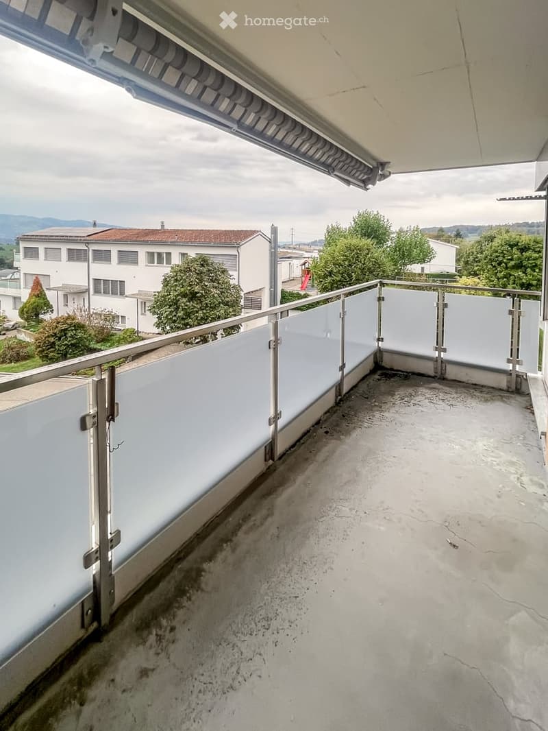 Balcony with modern glass railing, concrete floor, and a view of surrounding buildings and greenery.