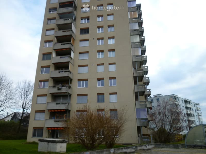 Tall residential building with multiple floors, balconies, and windows. Beige facade with brown window frames. Some balconies have railings and small awnings.