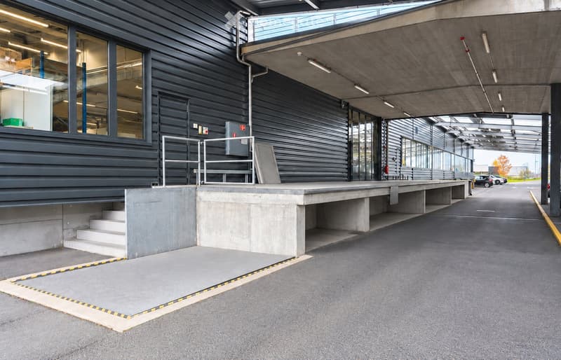 Modern building entrance with a raised ground floor, concrete platform, and glass walls. The entrance features a ramp and steps leading up to the building.
