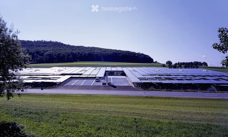 Large industrial building with solar panels on the roof, surrounded by open fields and a parking area.