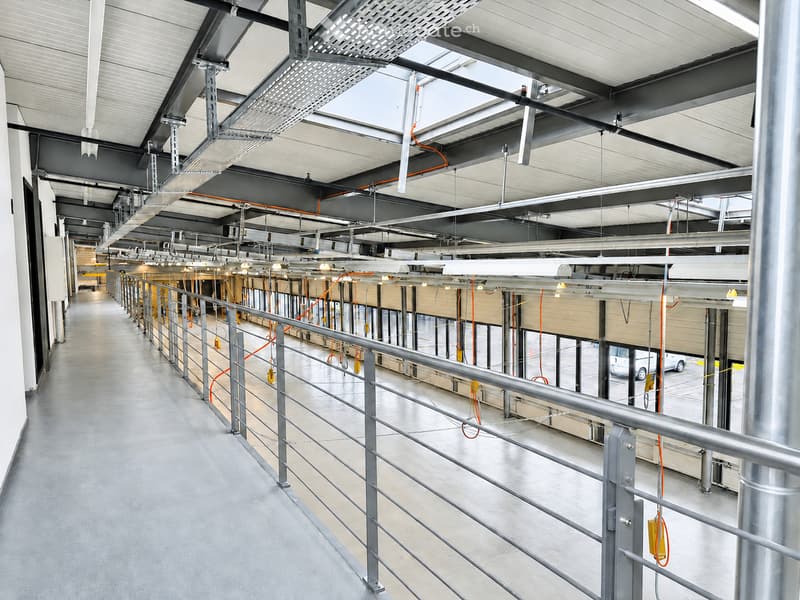 Modern industrial-style corridor with metal railings, exposed structural beams, and overhead lighting. The space features a polished concrete floor and visible electrical conduits.