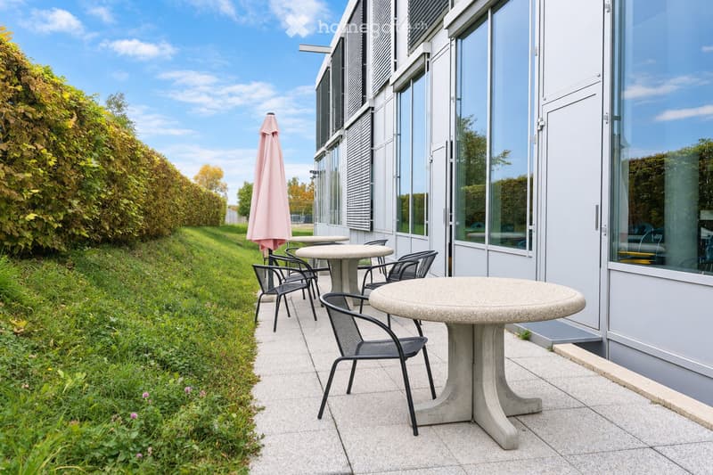 Outdoor seating area with tables, chairs, and an umbrella near a modern building with large windows and shutters.