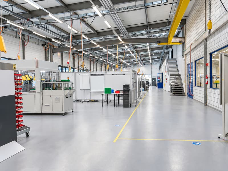 Modern industrial-style workspace with grey flooring, white walls, exposed metal beams, and bright overhead lighting. Includes workstations, tables, chairs, cabinets, and shelves.