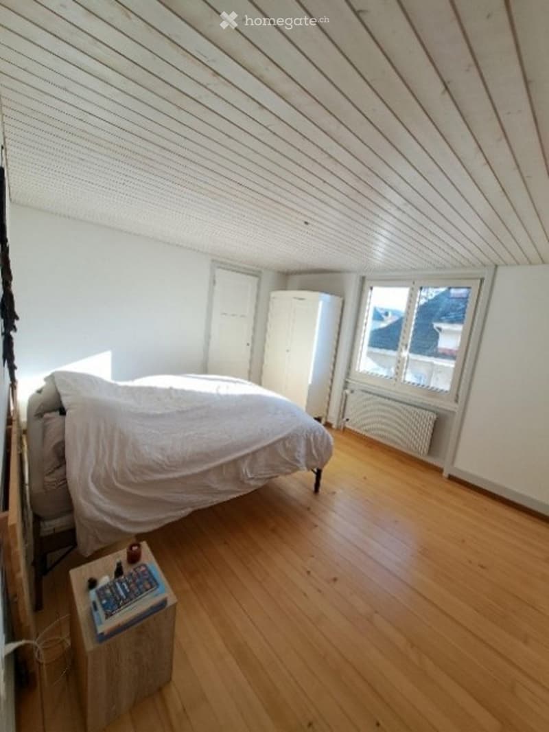 Bright bedroom with wooden flooring, slanted ceiling, large window offering a view, bed with white bedding, nightstand with books, and a wardrobe.