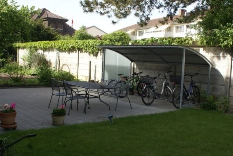 Outdoor patio area with a covered bike shelter, a table and chairs, and several bicycles. Potted plants are placed on the brick patio near the grassy area.
