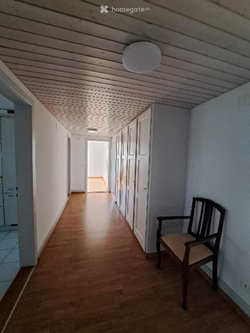 A narrow hallway with wooden flooring and white walls, featuring a single wooden chair placed against the wall.