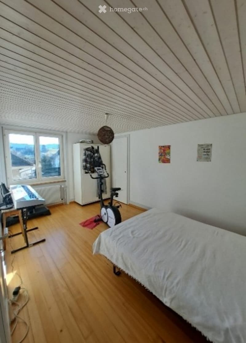 A multi-functional room with a bed, exercise bike, keyboard, and a cabinet. The room features wooden flooring, white walls, and large windows with a scenic view.
