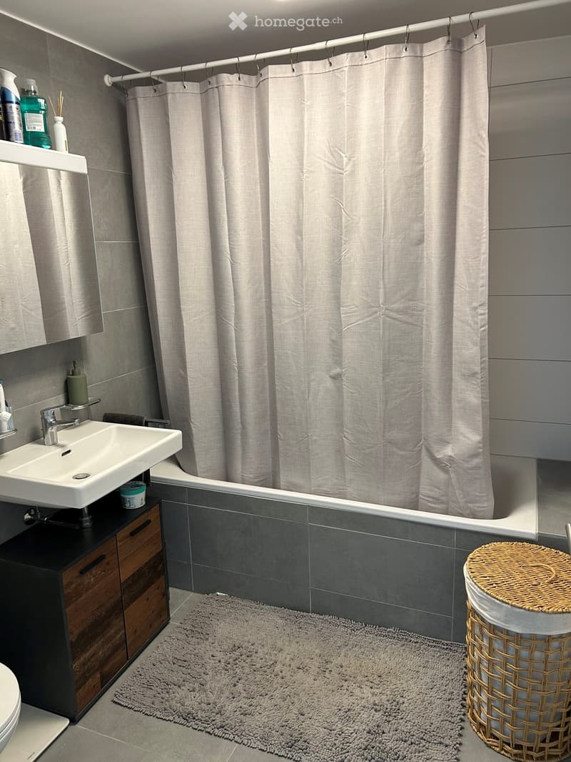 Modern bathroom with a white sink, chrome faucet, bathtub with a grey shower curtain, dark grey tiles, a woven stool, and a grey rug.