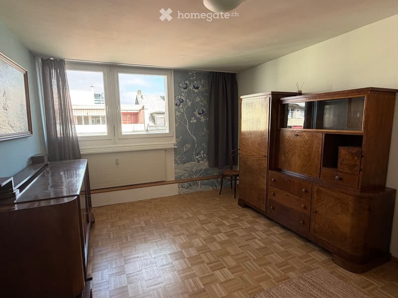 Empty room with wooden herringbone flooring, large window with floral wallpaper, wooden cabinet, and a small chair near the window.