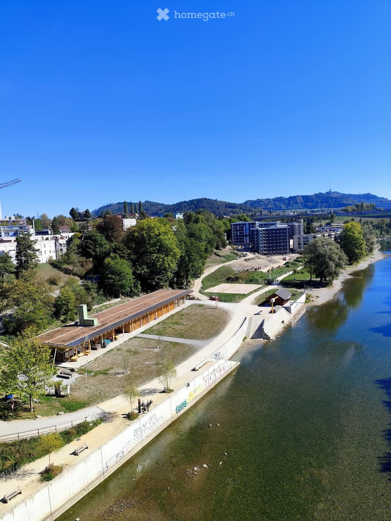 Aerial view of a riverside park area with pathways, benches, a small pavilion, and surrounding greenery, with a river flowing alongside.
