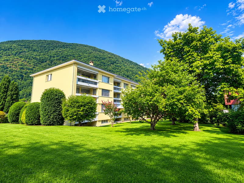 A yellow multi-story building with balconies, surrounded by a well-maintained lawn and trees, with a forested hill in the background.