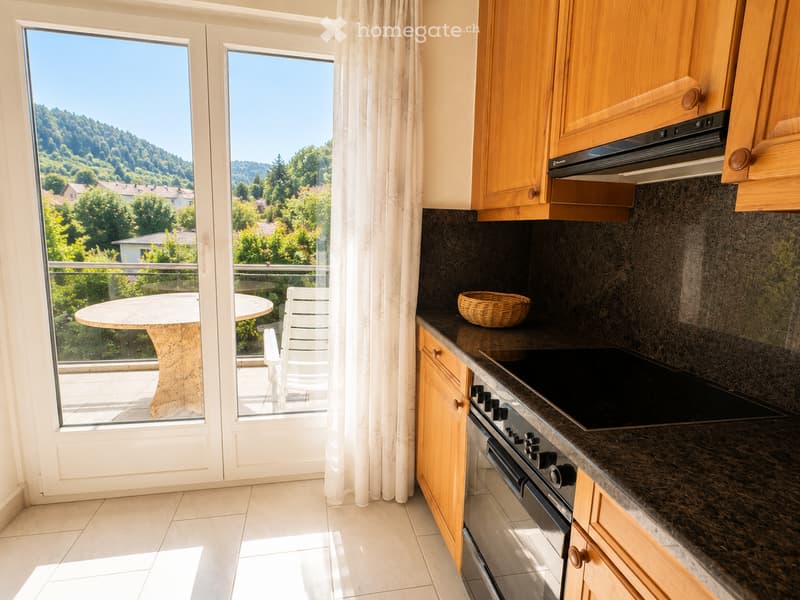Bright kitchen with wooden cabinets, granite countertops, and a view of a green landscape through large windows. Includes a round table and chairs on a balcony.