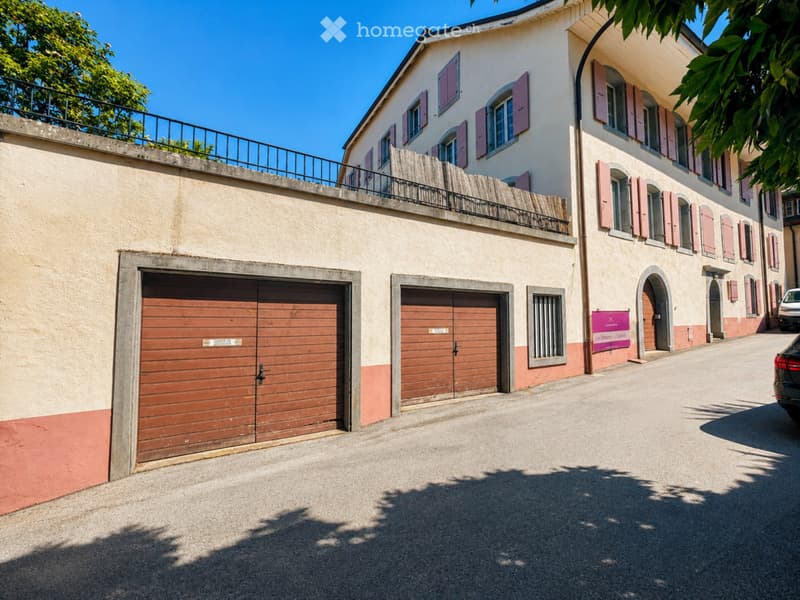 Two-story building with multiple garage doors, pink shutters, and a raised ground floor with a balcony.