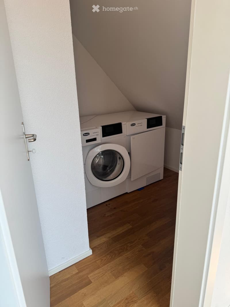 Small room with wooden flooring and white walls, featuring a washing machine and dryer unit.