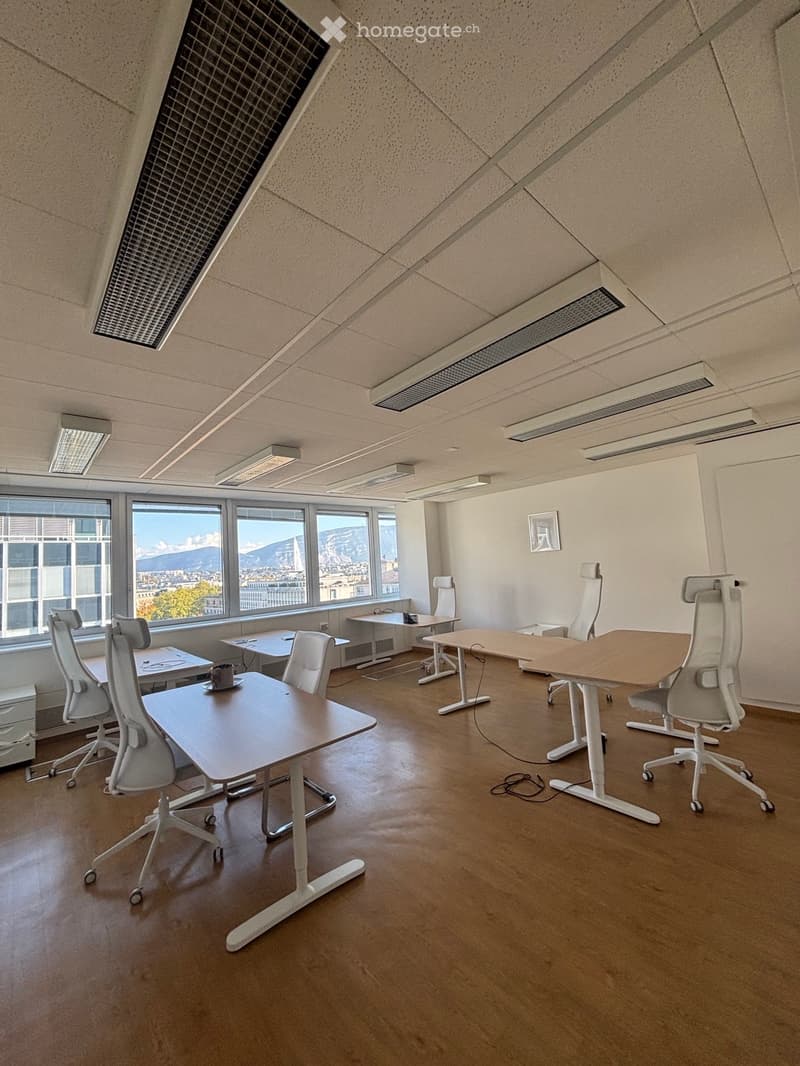 Modern office space with multiple desks, ergonomic chairs, and a cup on one of the desks. Large windows offer a scenic mountain view.