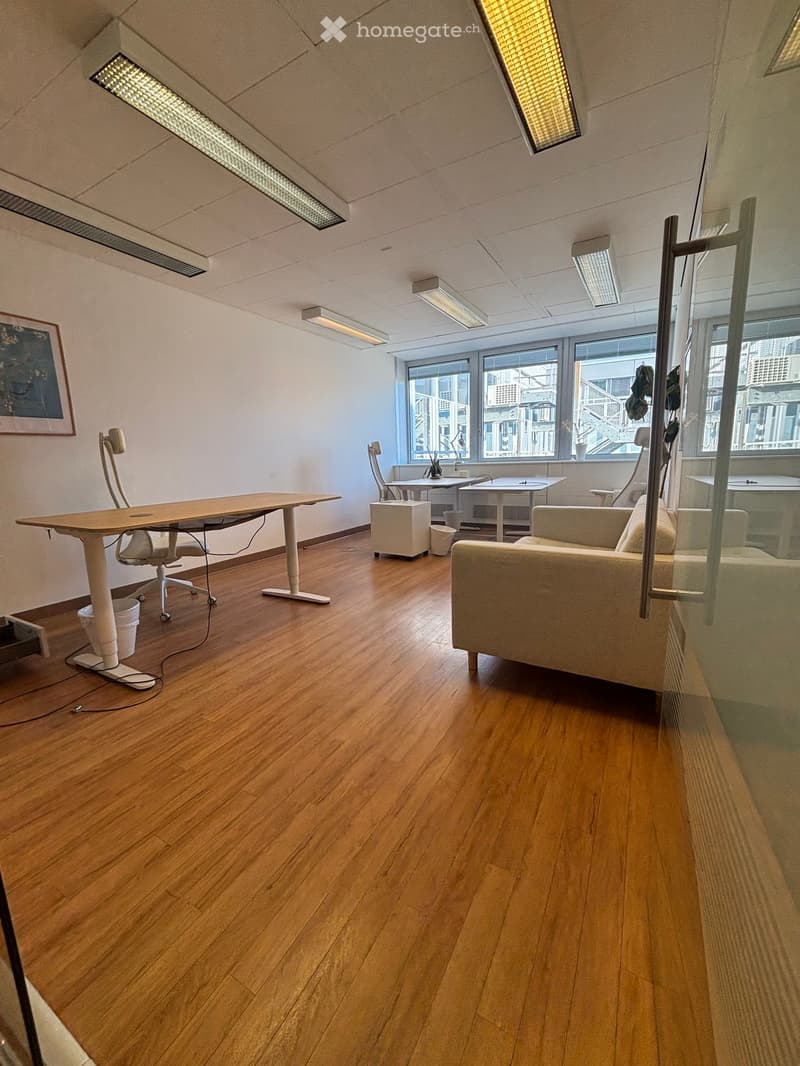 Modern office space with wooden flooring, large windows, a sofa, office chair, desk, and a screen.