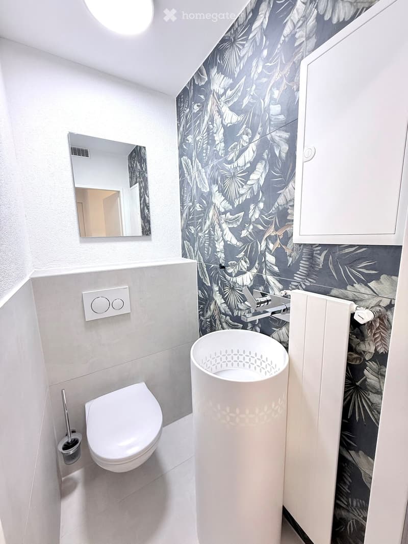 Modern bathroom with a toilet, sink, mirror, and cabinet. The walls feature a leafy pattern, and the sink has a sleek, contemporary design.