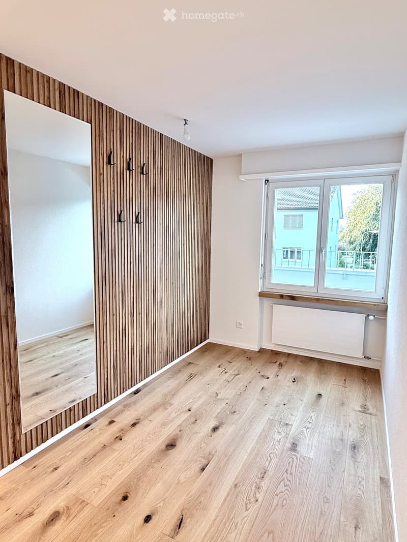 Empty room with light wooden flooring, white walls, and a large window with a view of a neighboring building. The room features a radiator and a coat rack with hooks.