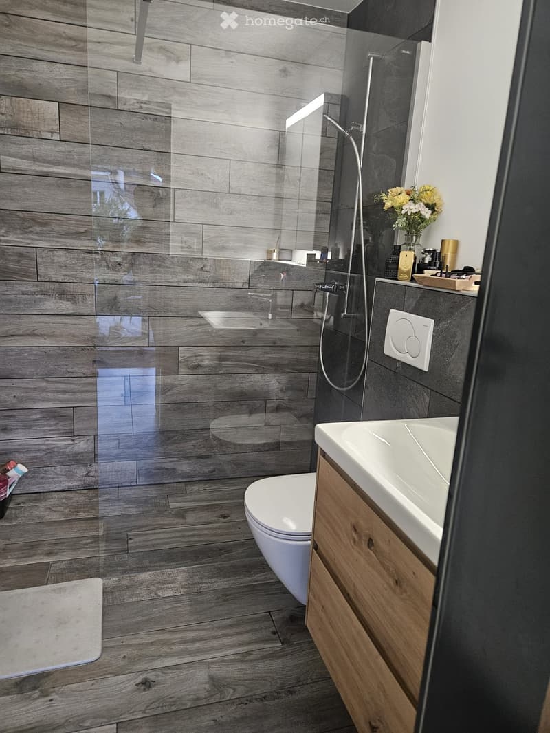 Modern bathroom with wooden flooring, a white toilet, a wooden vanity with a sink, a glass-enclosed shower, and a vase with flowers on the counter.