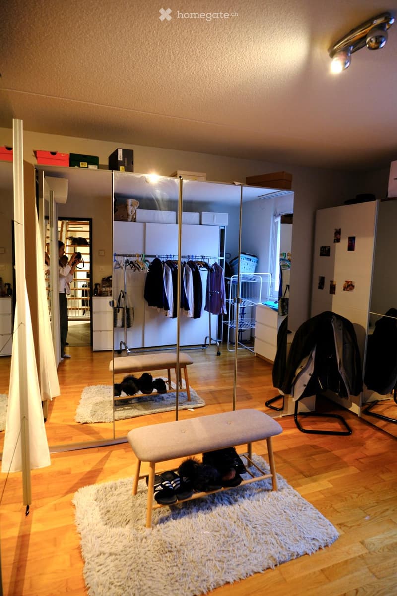 A well-lit, organized walk-in closet with wooden flooring, a large mirror, a clothing rack, a shoe bench, and a rug.