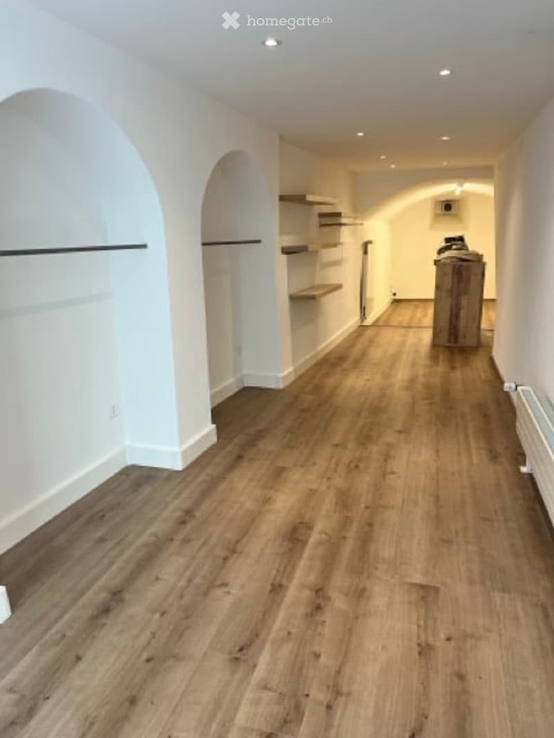 Empty room with hardwood floors, white walls, arched doorways, wooden shelves, power outlet, wooden stool, radiator.