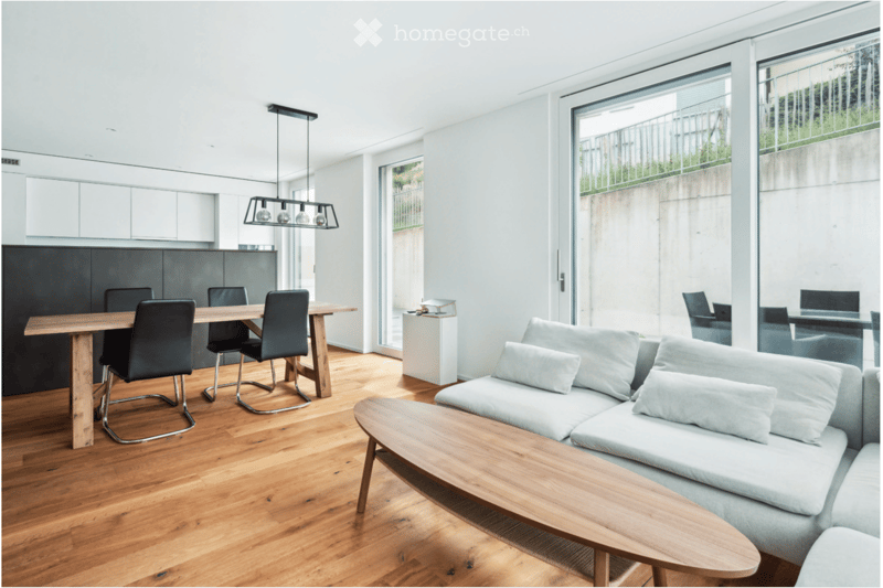Open-plan living and dining area with light grey sofa, wooden coffee table, dining table with chairs, pendant lighting, and modern kitchen cabinets.