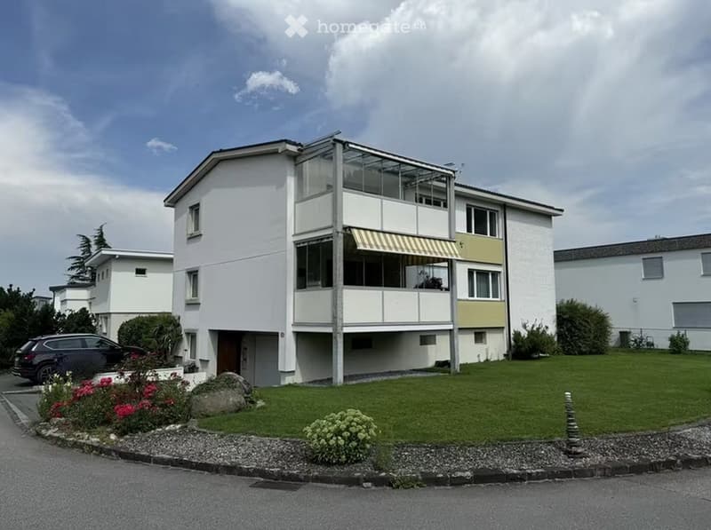 Modern two-story house with white exterior, large windows, and a balcony on the upper floor. The house features a well-maintained lawn, colorful flowers, and a parked car in the driveway.