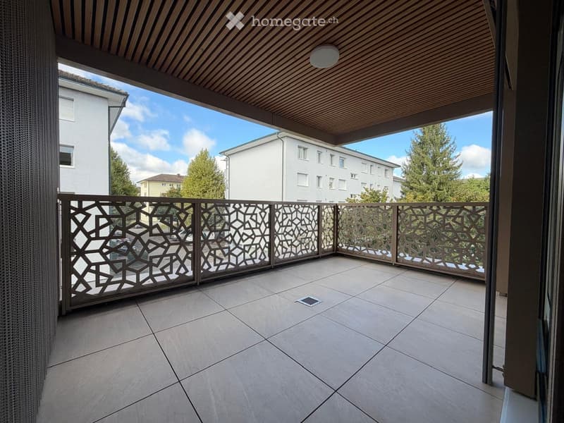 Balcony with tiled floor, decorative metal railing, wooden ceiling, and view of surrounding buildings and trees.