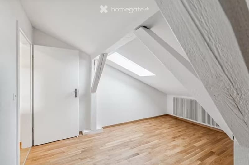Bright attic room with wooden flooring, white walls, exposed wooden beams, and skylight windows. No furniture or equipment present.
