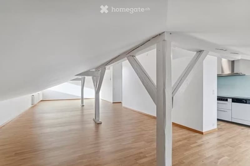 Open, loft-style room with wooden flooring, white walls, exposed wooden beams, and a small kitchen area with white cabinets and stainless steel appliances.