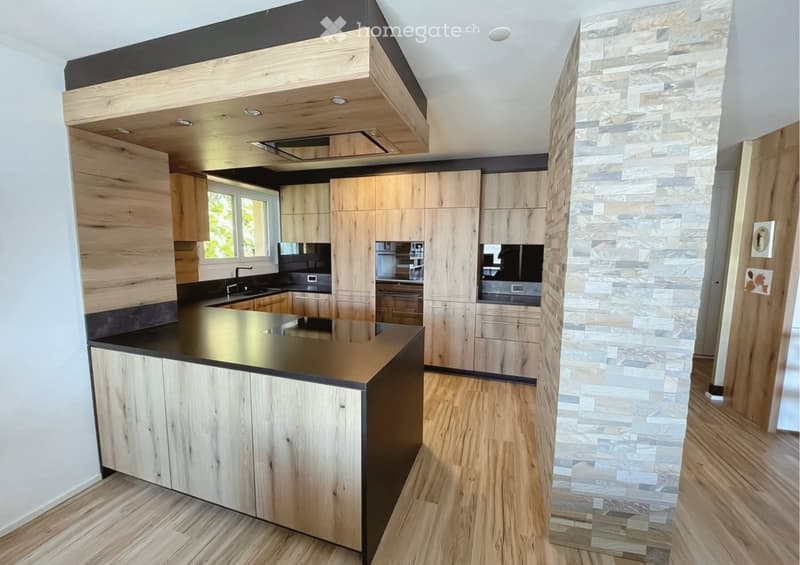 Modern kitchen with wooden cabinets, a central island, and a stone accent wall. Features include a black countertop, a sink, and a window providing natural light.