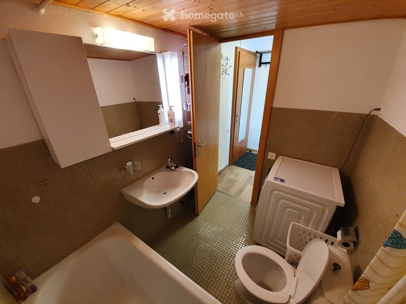 Bathroom with a sink, toilet, bathtub, mirror, and washing machine. The room has tiled walls and a wooden ceiling.
