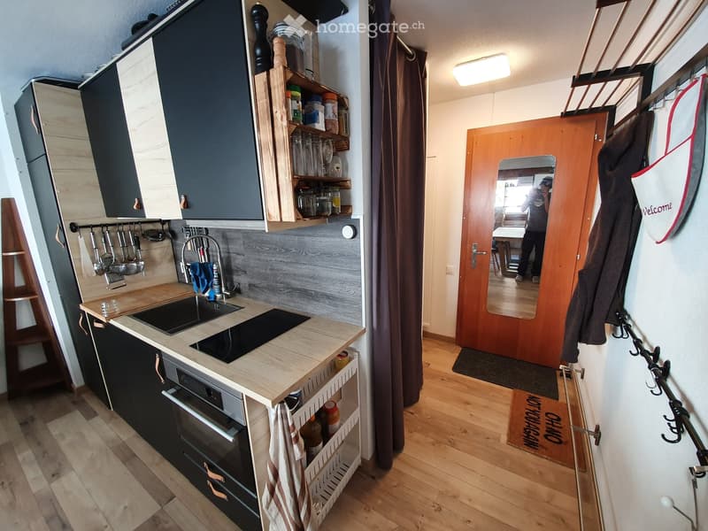 Modern kitchen with wooden countertops, black cabinets, sink, oven, and shelves holding jars and glasses. Wooden floor and a hallway with a coat rack and a welcome sign are visible.