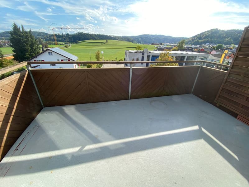 Empty wooden balcony with a metal railing, overlooking a green landscape and town with houses and trees.