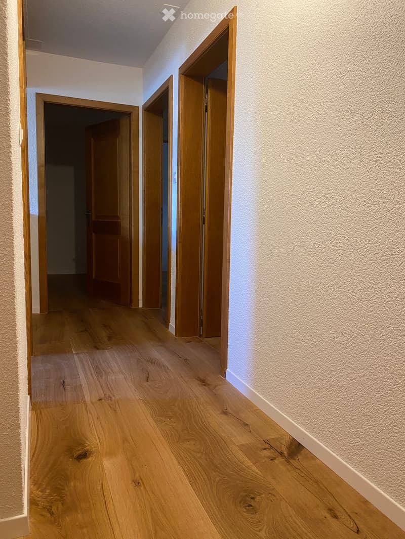 A clean, well-lit hallway with wooden flooring and white walls, featuring several open wooden doors leading to other rooms.
