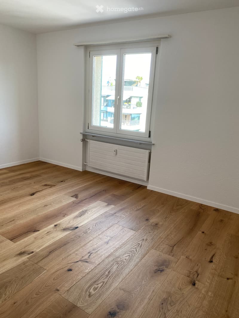 Empty room with light wooden flooring, white walls, and a large window with a radiator below it.