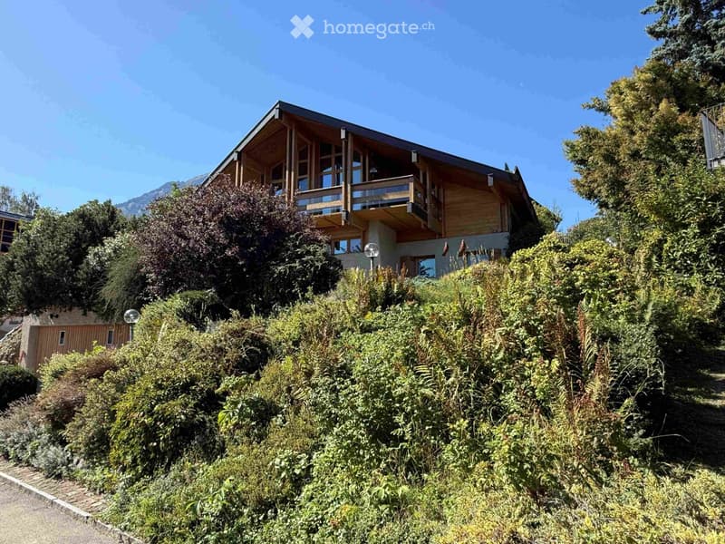 A two-story wooden house with large windows and a balcony, surrounded by lush greenery and trees.