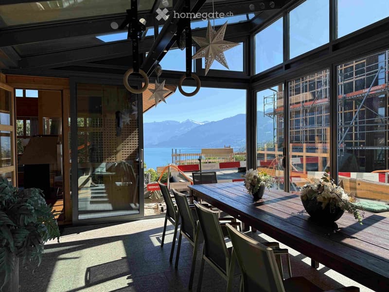 Bright, airy room with large windows offering a scenic mountain and lake view. Features a wooden dining table with chairs, flower vases, and a fireplace.